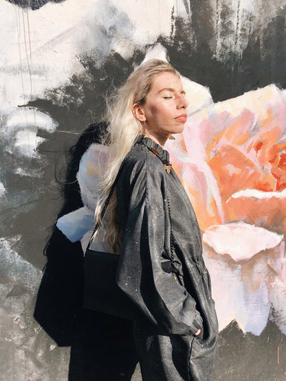 A woman with long blonde hair stands in front of a wall with a flower painting. Alt text: Chain Shoulder Bag - Black exudes elegance with genuine leather, sleek chain strap, flap front, and handcrafted European quality.