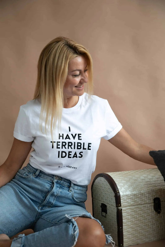 A woman sitting on a bench, wearing an oversized t-shirt with the text 'I Have Terrible Ideas'. Product made of organic cotton, featuring a simple message. Size chart available for XS to XL.