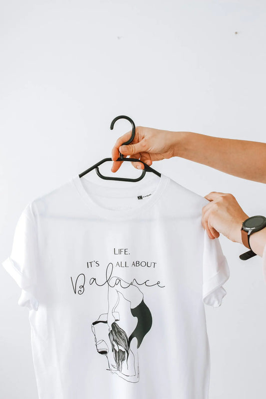 Person holding an oversized white 'Life. It's All About Balance' t-shirt on a hanger. Made of organic cotton. Size chart: XS to XL. Washing instructions included.