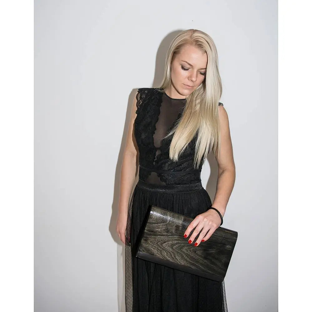 A woman in a black dress holds a Large Glossy Wood Clutch, showcasing leather and matte black wood. Handmade with a detachable shoulder strap, magnetic clasp closure, and interior zip pocket.