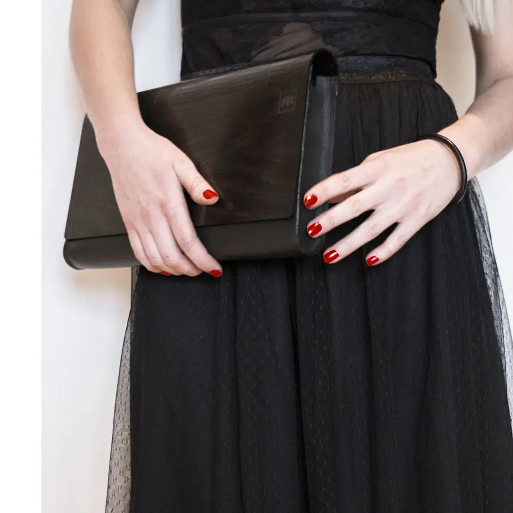 A woman holds a black wood and leather clutch bag with a detachable shoulder strap. Handmade from birch wood, each panel unique. Features magnetic clasp, interior zip pocket, versatile styling options.