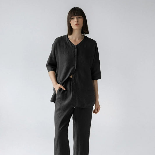 A woman in black linen Primrose loungewear set, featuring a V-neck top with 3/4-length sleeves and buttoned closure, paired with relaxed-fit trousers and an elastic waistband.