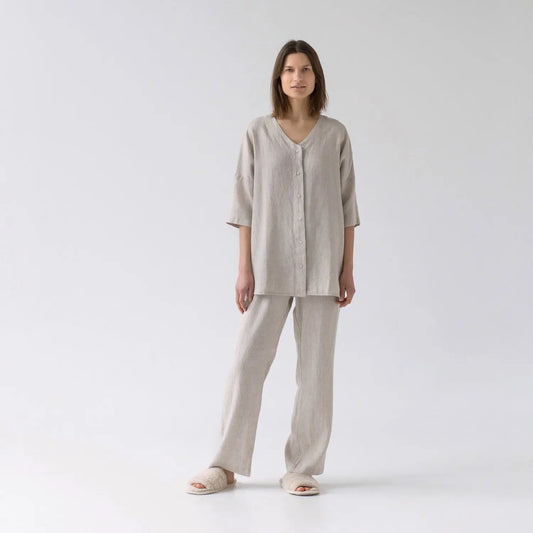 A woman in a white linen Primrose loungewear set, featuring a V-neck top with buttoned front closure and 3/4-length sleeves, paired with relaxed-fit trousers and an elastic waistband.