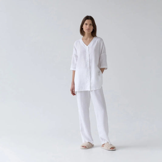 A woman in white linen Primrose loungewear set, featuring a V-neck shirt with 3/4-length sleeves and full buttoned closure, paired with relaxed-fit trousers and an elastic waistband.