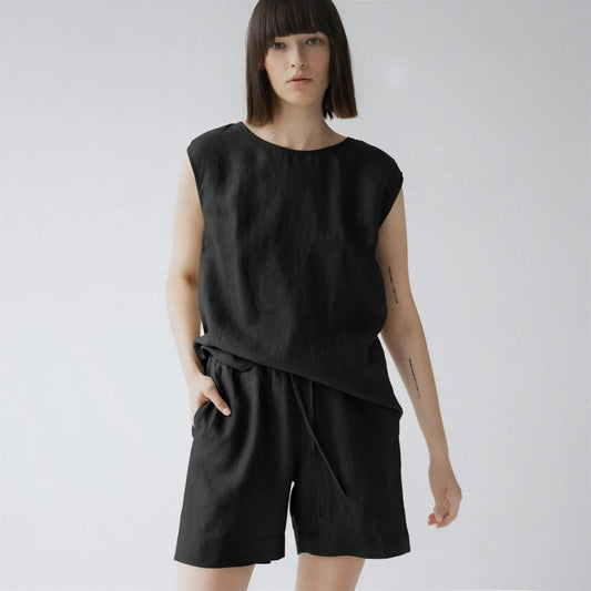 A woman in a black linen Verbena pyjama set, featuring a relaxed fit top with a round neck and sleeveless design, paired with trousers having an elasticated waistband and slanted side pockets.