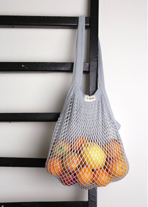 A plastic-free net bag hanging on a wall, filled with oranges. Versatile for shopping, gym, and beach. Made of 100% organic cotton, with strong handles and visible contents.