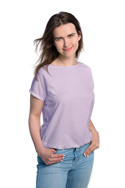 A woman in a tan-through lilac T-shirt, hands in pockets. Spacious cut for comfort during outdoor activities. Fabric allows healthy tanning with UVA ray penetration. Sizes XS-XXL available.