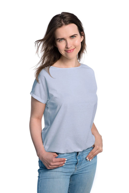 A woman in a light blue Tan-Through T-shirt, hands in pockets, showcasing its spacious cut for outdoor comfort and UV protection fabric blend.
