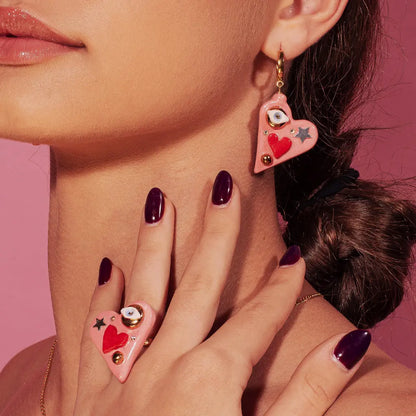 Close-up of woman's ear wearing heart-shaped earrings with Eye of Protection charm, SWAROVSKI crystal, and 24K gold details. Hand-made ceramic earrings, unique variations.