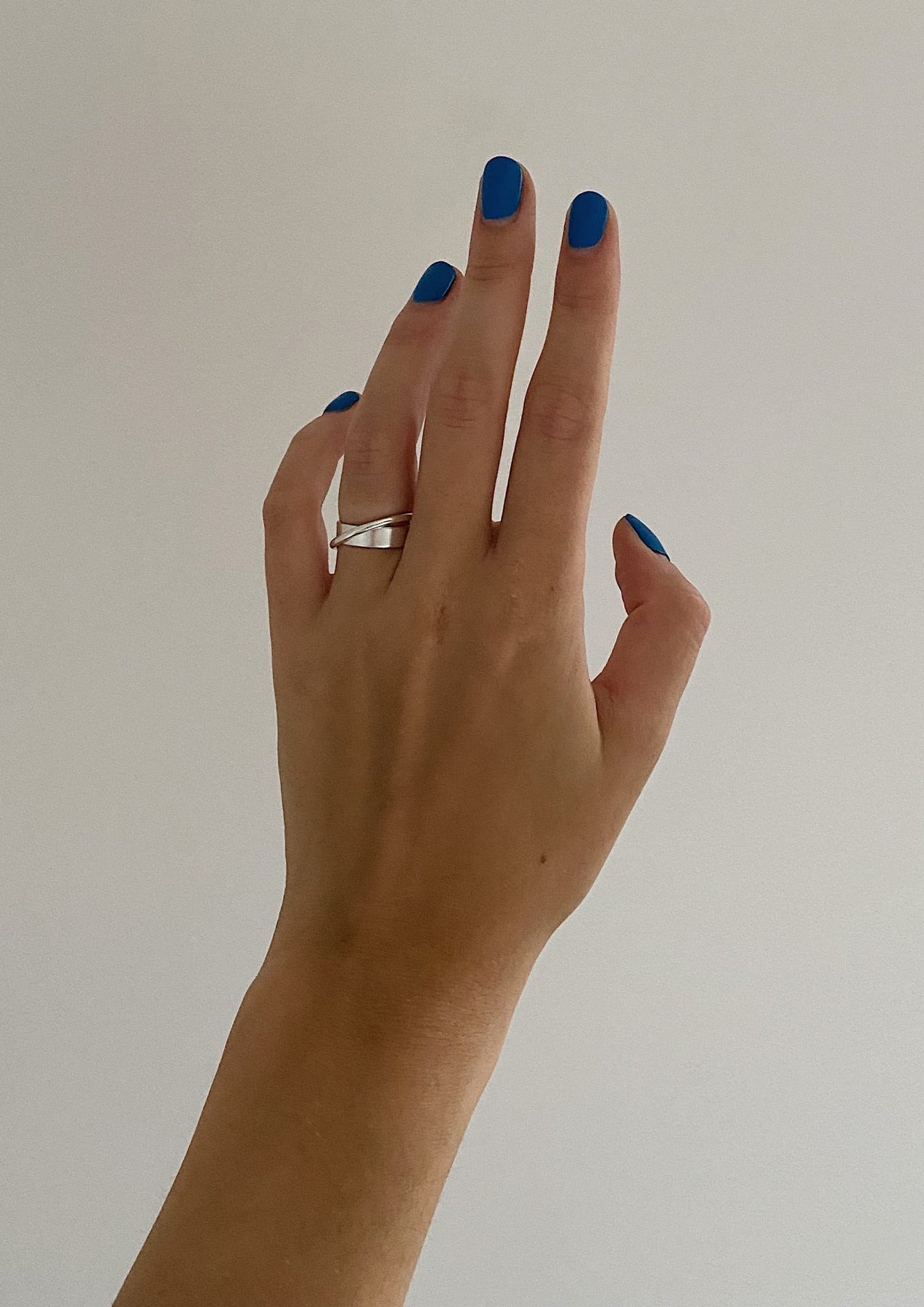 A hand wearing the Andromeda Ring - Silver, showcasing blue painted nails. Hand-crafted sterling silver ring with unique angles, 3mm thick, 7.3mm high, 4.1-4.5g weight. Sustainable packaging. Made in Europe.