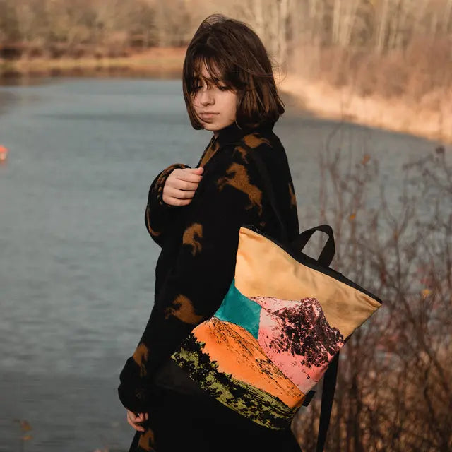 A woman in a black and brown coat stands near water, holding a backpack featuring a mountain landscape design. This spacious velvet and polyester backpack includes adjustable straps and pockets for a 13 laptop.