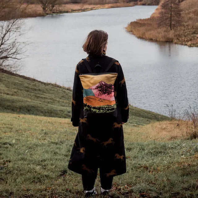 A person stands by a river on grass, wearing a black and brown dress. A pillow with a mountain image. Alt text: Backpack - Bold Mountains, velvet top, waterproof polyester bottom, laptop pocket, adjustable straps.