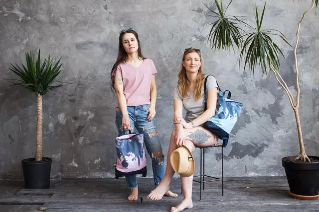 Two women posing with a PINK “Watercolor” backpack featuring velvet top, waterproof navy blue bottom, and blue lining. Adjustable straps, zipper closure, and laptop pocket included.