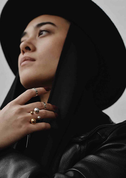 A woman wearing a black hat and jacket, showcasing the Big Bomb Multisize Ring - Gold. Handmade in Lithuania, this statement ring features 6mm and 10mm bubbles on a 1.8mm band.