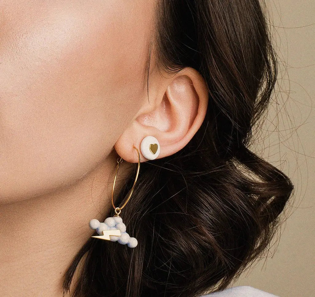 Close-up of Eye in the Sky earrings on a woman's ear, showcasing a heart-shaped charm and gold hoop detail. Hand-made ceramic with 24K gold lustre and protective symbolism.
