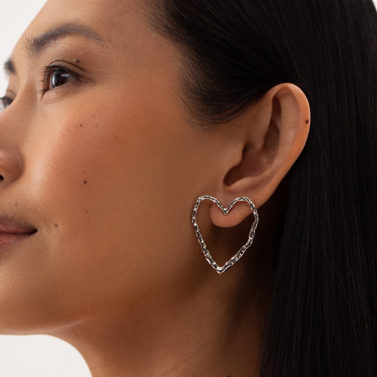 A close-up of a woman wearing minimalist heart-shaped Eros silver stud earrings. Sustainable, hypoallergenic, and easy to wear for all-day comfort.