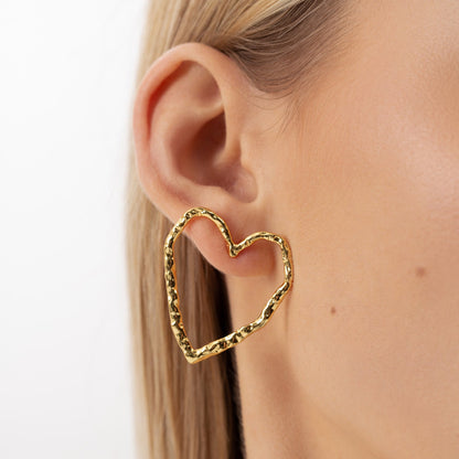 Close-up of a woman's ear wearing minimalist heart-shaped Eros silver stud earrings. Sustainable, hypoallergenic, and elegant. Made from recycled silver, 18k gold plated.