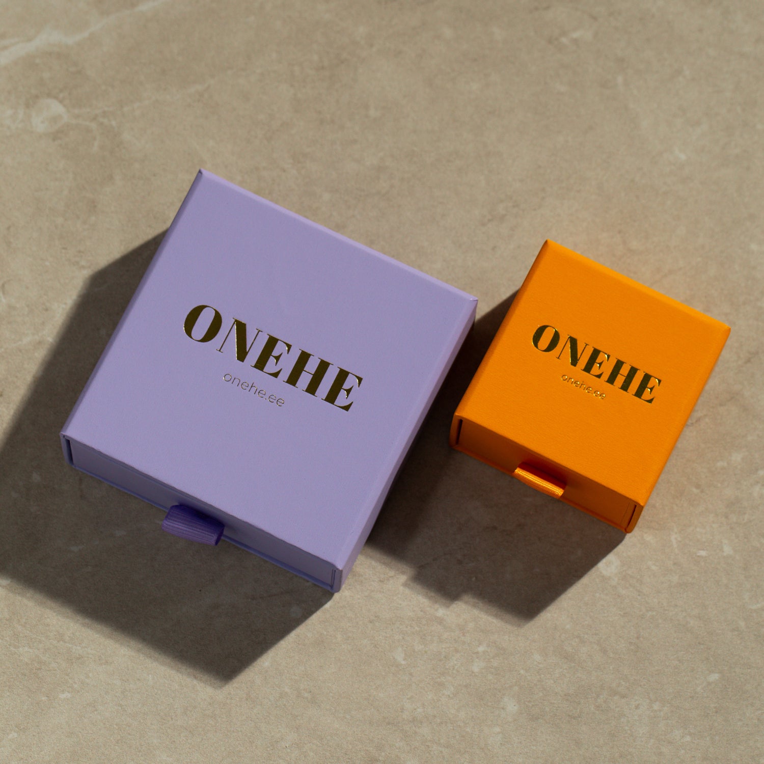 Golden heart-shaped stud earrings on marble surface, purple and orange boxes with gold text, close-up of logo. Sustainable, hypoallergenic ONEHE earrings made from recycled silver, plated with 18k gold. Size: 7 x 6.5 mm.