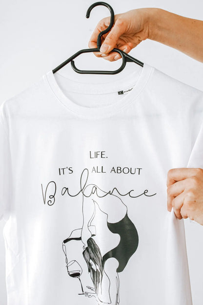A person holding an oversized white shirt with the print Life. It's All About Balance. Made of organic cotton. Includes size chart and washing instructions.