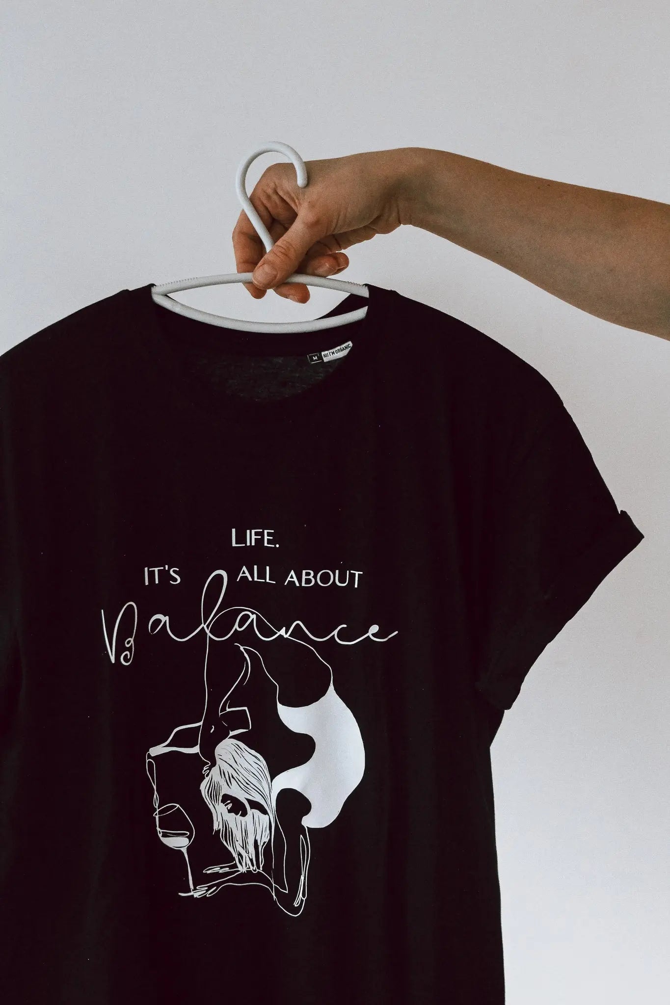 A black shirt held in a hand, featuring 'Life. it's all about balance' print. Oversized organic cotton t-shirt with size chart and washing instructions.