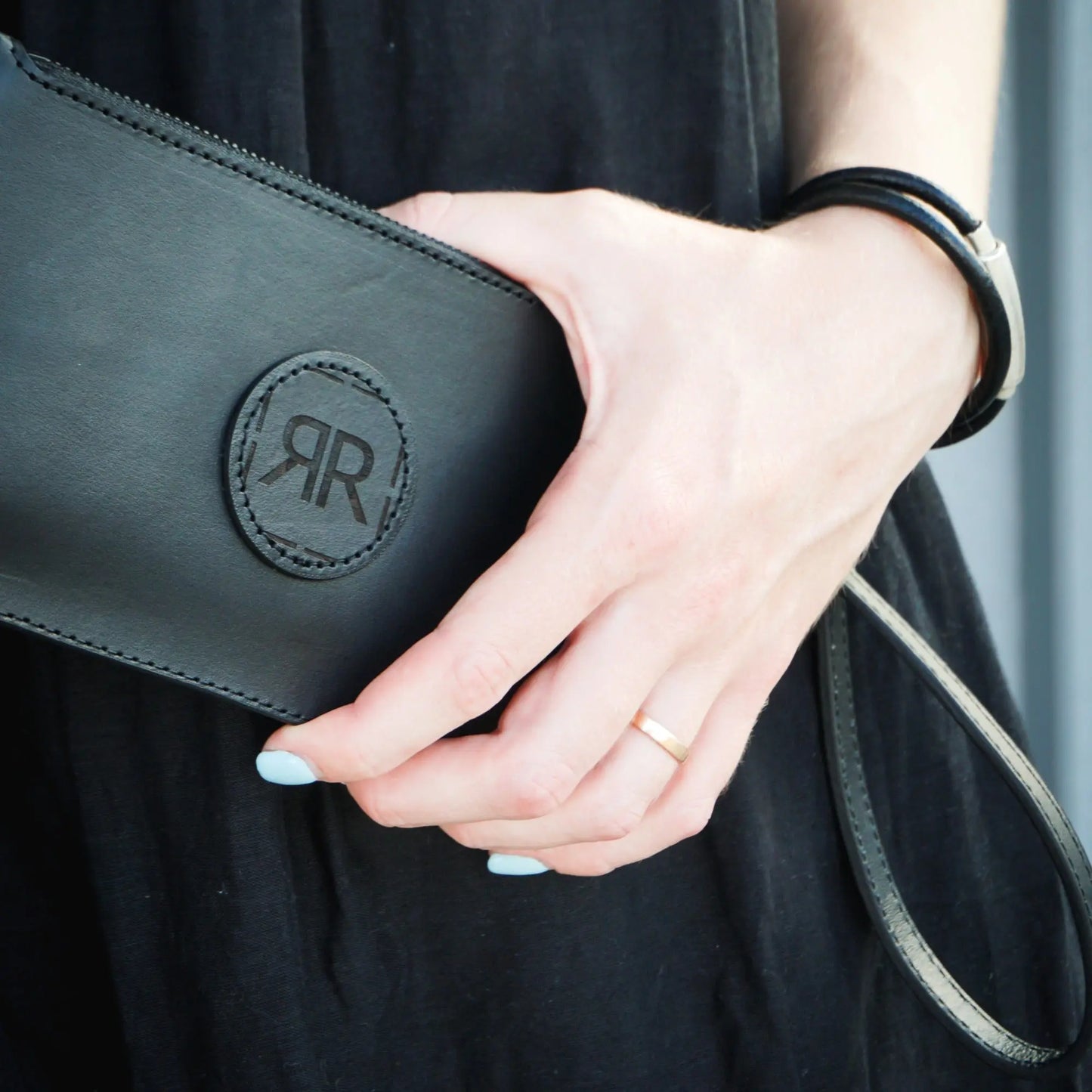A person holding a black purse with a removable leather wristlet, featuring a logo detail and compartments for cards and cash.