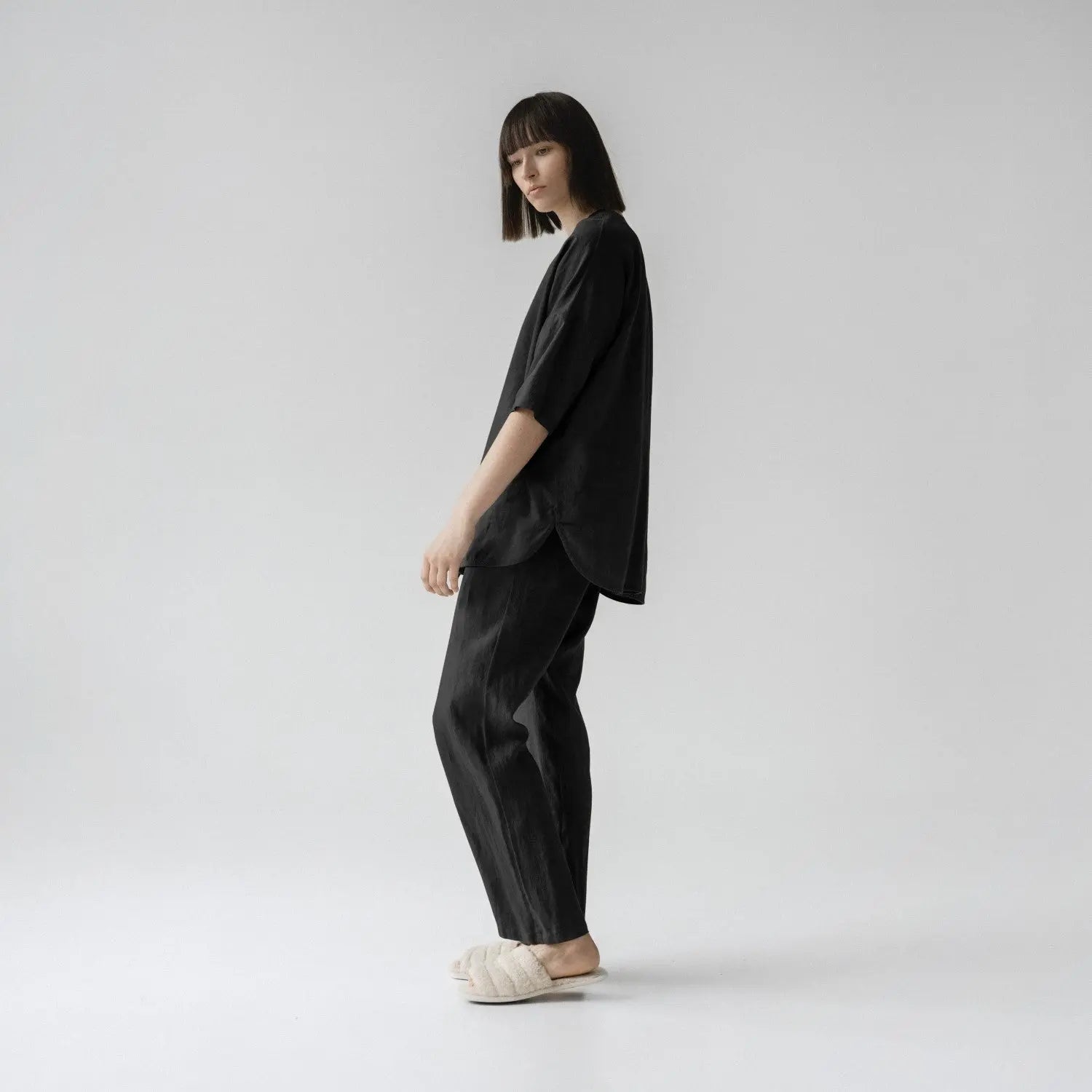 A woman in black linen Primrose loungewear set, featuring a relaxed fit shirt with V-neck and buttoned closure, paired with trousers with an elastic waistband and side pockets.