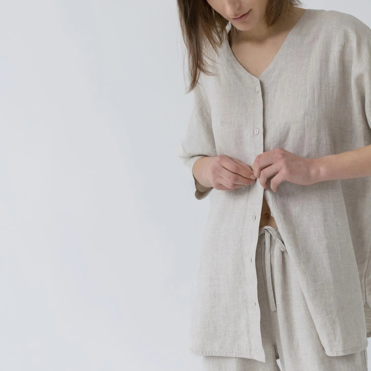 A woman buttoning a linen Primrose loungewear set, featuring a V-neck top with buttoned front closure and 3/4-length sleeves, paired with relaxed-fit trousers and a matching linen bag.