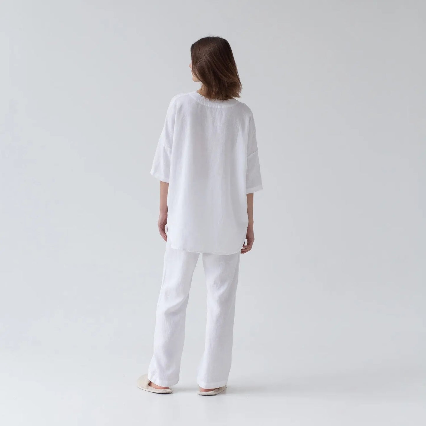 A woman in white linen Primrose loungewear set, featuring a V-neck shirt with 3/4-length sleeves and buttoned front closure, paired with relaxed-fit trousers and an elastic waistband.
