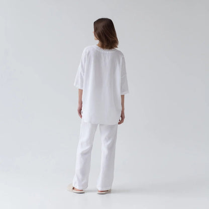 A woman in white linen Primrose loungewear set, featuring a V-neck shirt with 3/4-length sleeves and buttoned front closure, paired with relaxed-fit trousers and an elastic waistband.