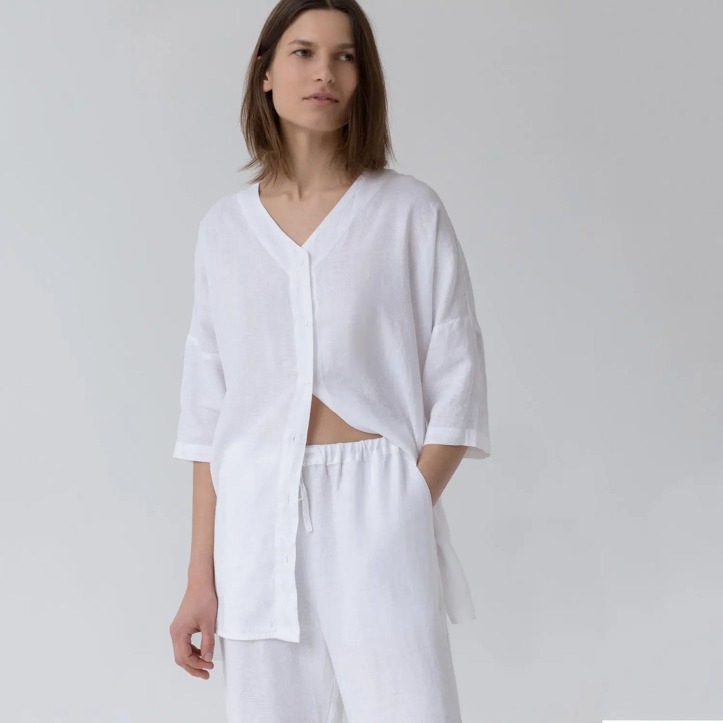 A woman in white Linen Primrose loungewear set, featuring a V-neck shirt with 3/4-length sleeves and buttoned front, paired with relaxed fit trousers and an elastic waistband.