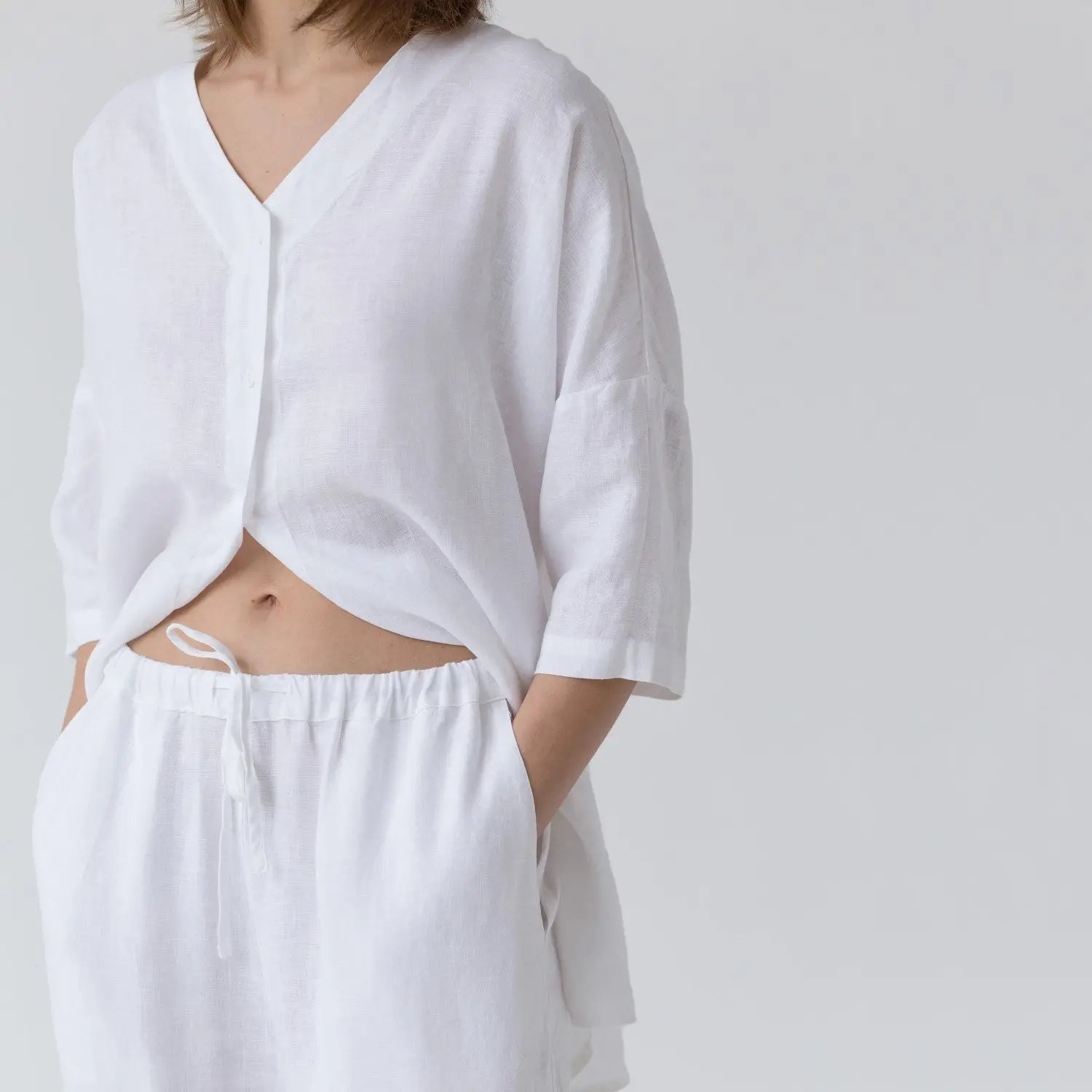 A woman in white linen Primrose loungewear set, featuring a V-neck shirt with buttoned front and 3/4 sleeves, paired with relaxed-fit trousers and slanted pockets. Handmade from 100% linen, perfect for lounging.