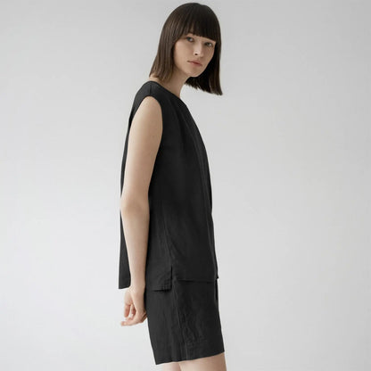 A woman in a black linen Verbena pyjama set, featuring a relaxed fit top with a round neck and sleeveless design, paired with trousers having an elasticated waistband and slanted pockets.