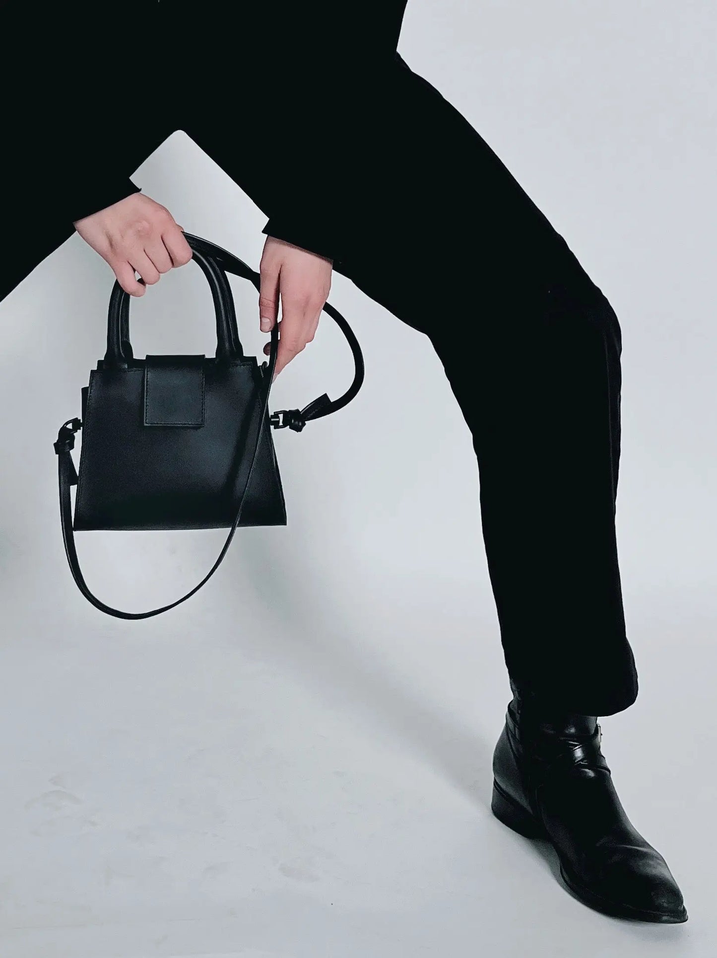 A person holds a black Mini Leather Handbag with reinforced handles, zipper closure, and detachable shoulder strap. Crafted from high-quality leather, featuring knot detailing and interior pocket for organization.