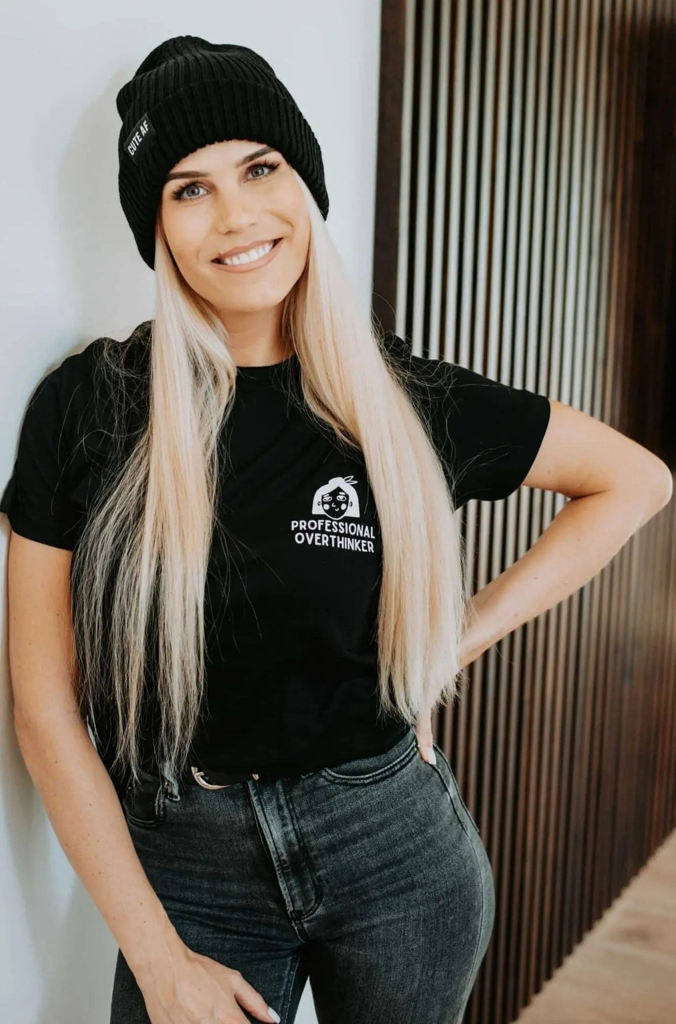 A woman in a black hat and shirt, featuring a close-up of a cartoon face. Product: Professional Overthinker T-shirt, oversized organic cotton with a printed message. Size chart included.
