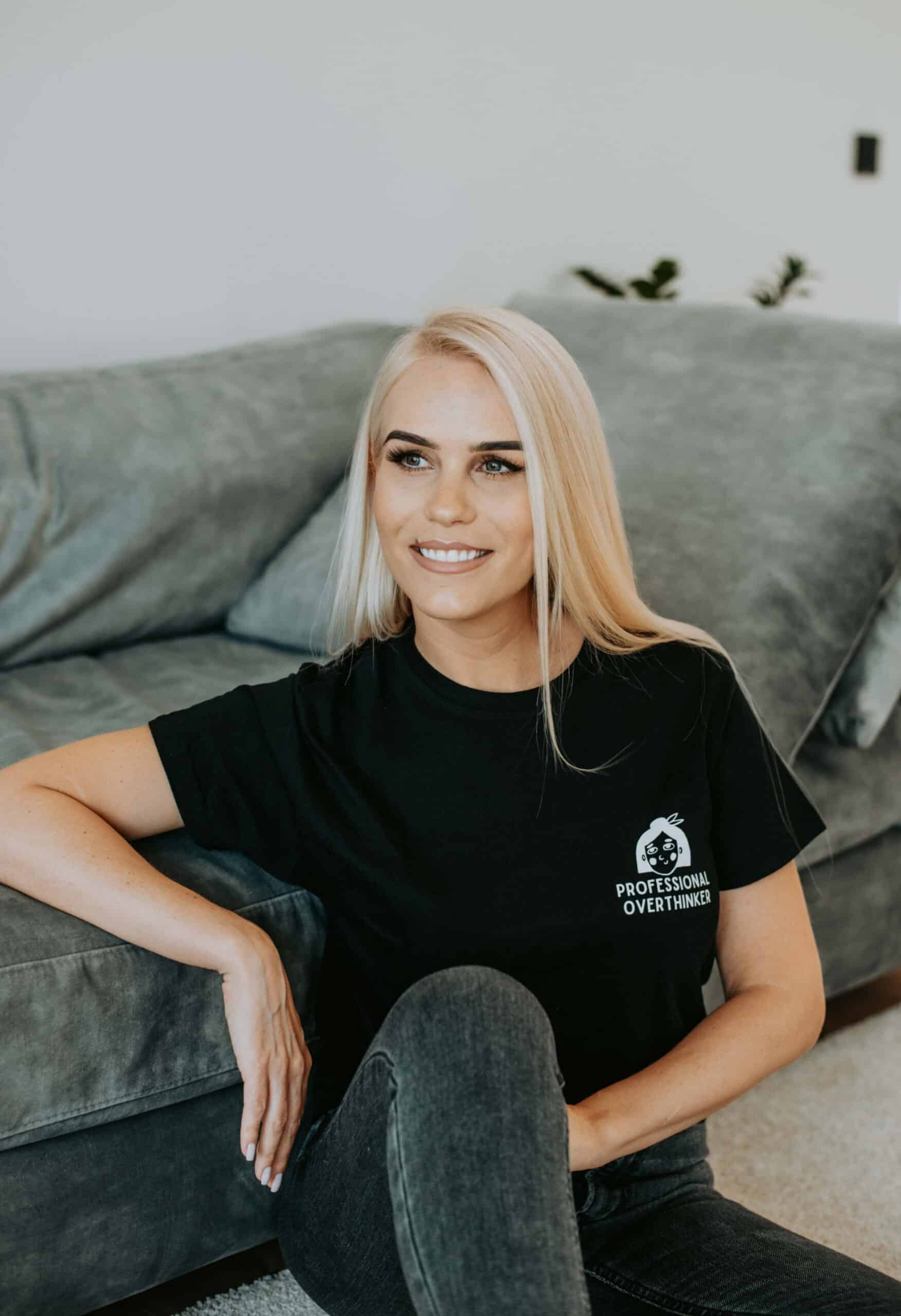 A woman in a black shirt sits on a couch, smiling at the camera. She wears an oversized organic cotton t-shirt with Professional Overthinker printed on it.