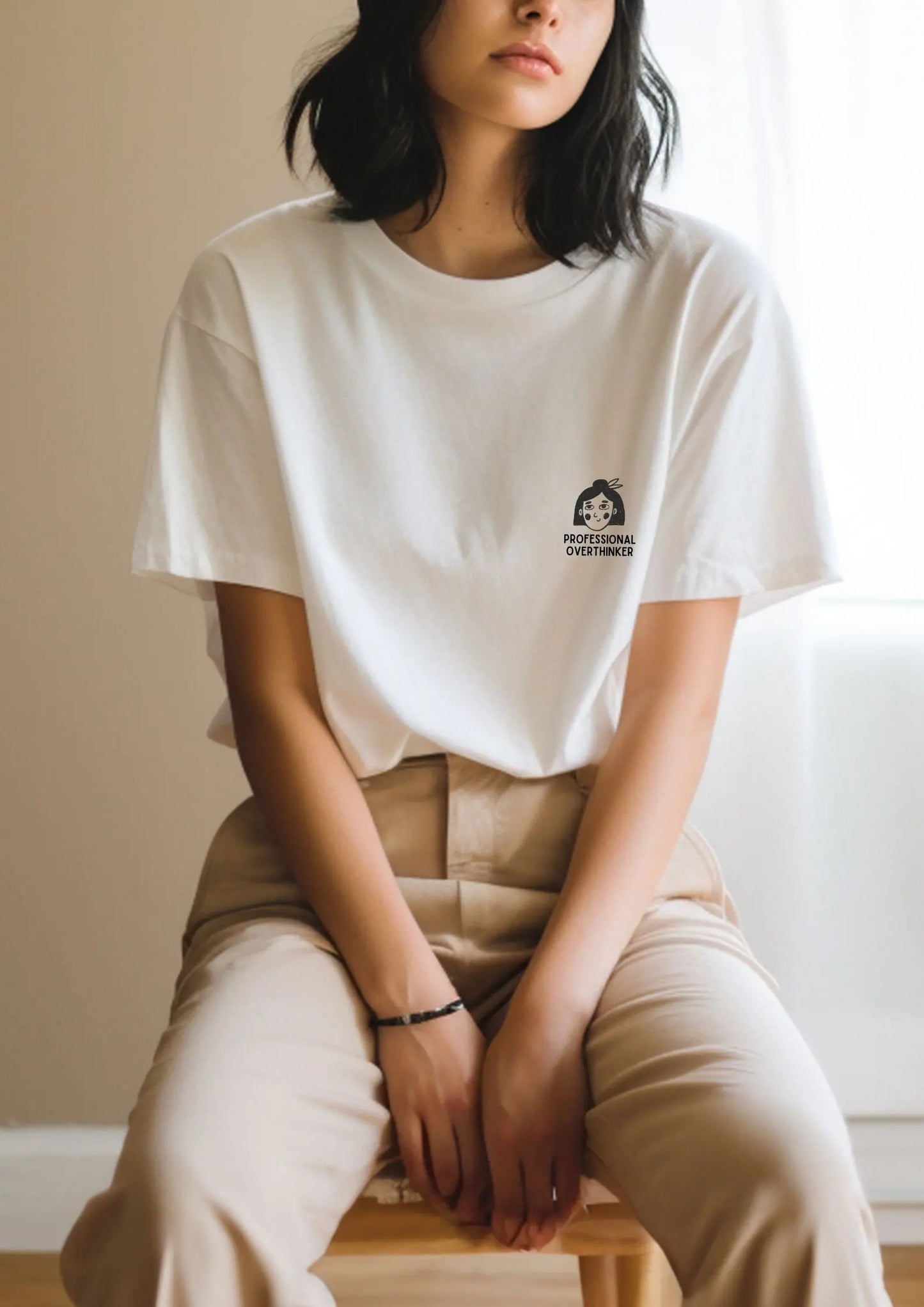 A woman in a white shirt sits in a chair, showcasing an oversized organic cotton T-shirt with Professional Overthinker print. Size chart and washing instructions included.