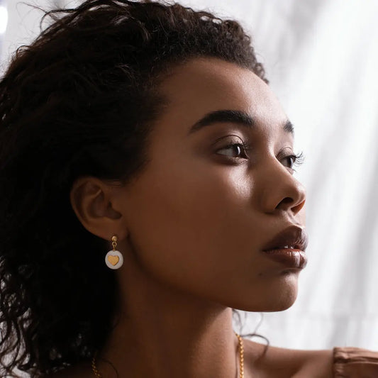 A woman with a heart-shaped gold pendant, showcasing Pure Love earrings. Hand-made ceramic pendants with 24K gold-over 925 sterling silver details. Unique variations possible.