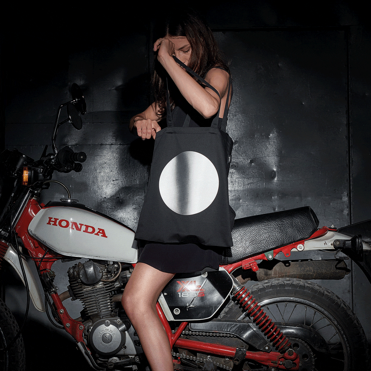 A woman stands on a motorcycle, holding a Reflective Tote DOT by MARCH Design Studio. The tote features a reflective print, inside pocket, and European craftsmanship.