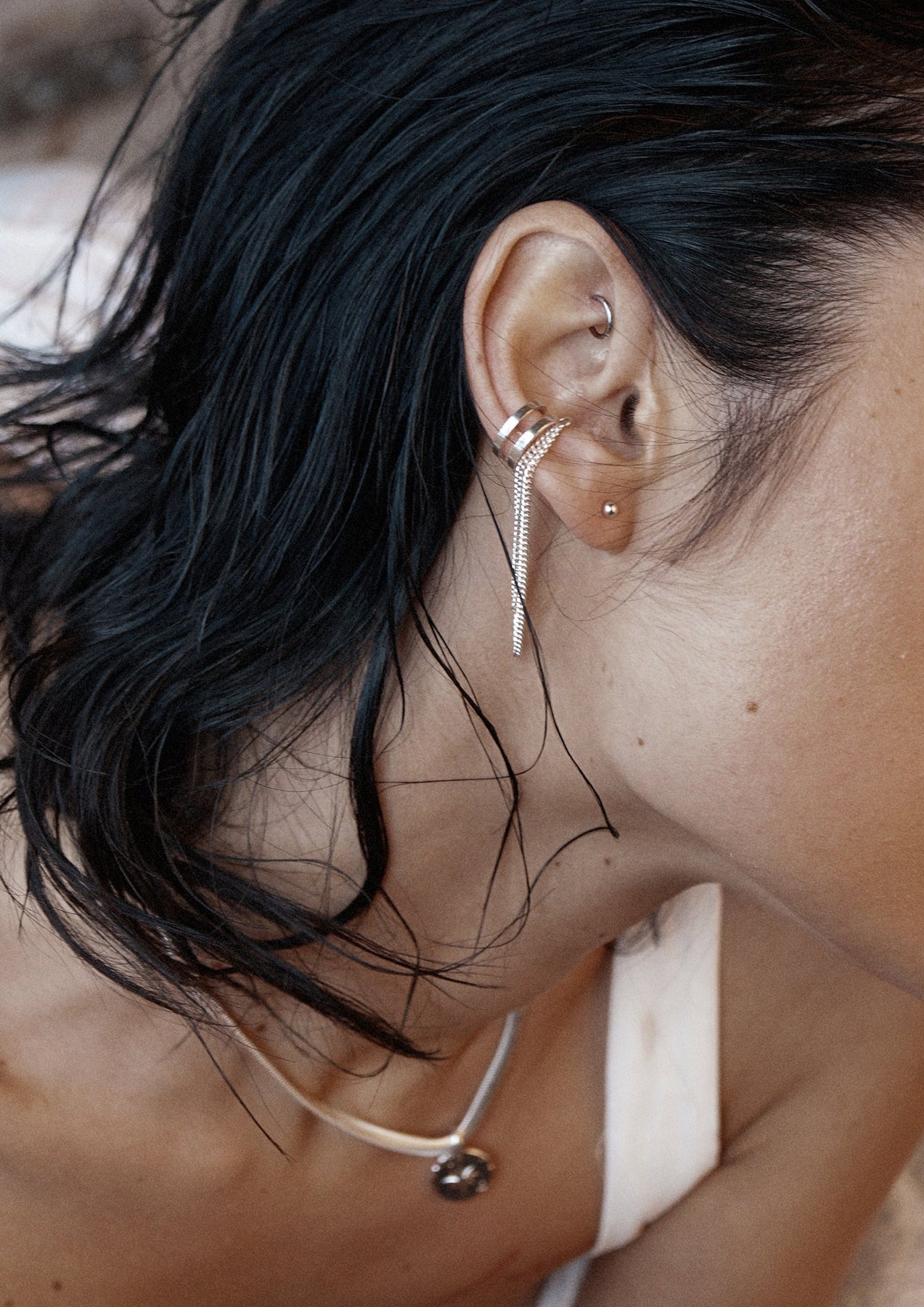 A close-up of a woman wearing the Revolve Ear Cuff in sterling silver, featuring a double chain design. Handcrafted without the need for ear piercings. Dimensions: 1mm thickness, 5.73mm width, 60mm length.