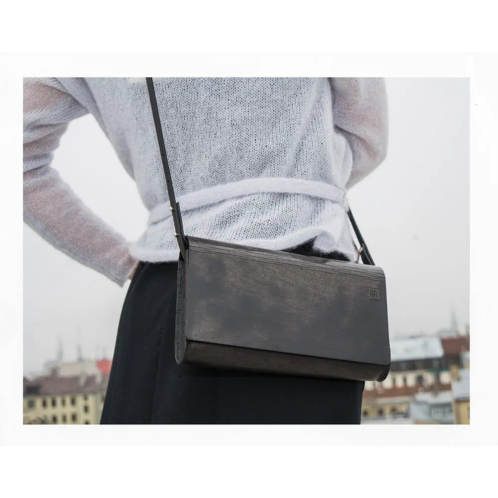 A person holding a Small Glossy Wood Clutch bag with detachable shoulder strap, showcasing leather body and matte black wood panel. Handmade from birch tree wood.