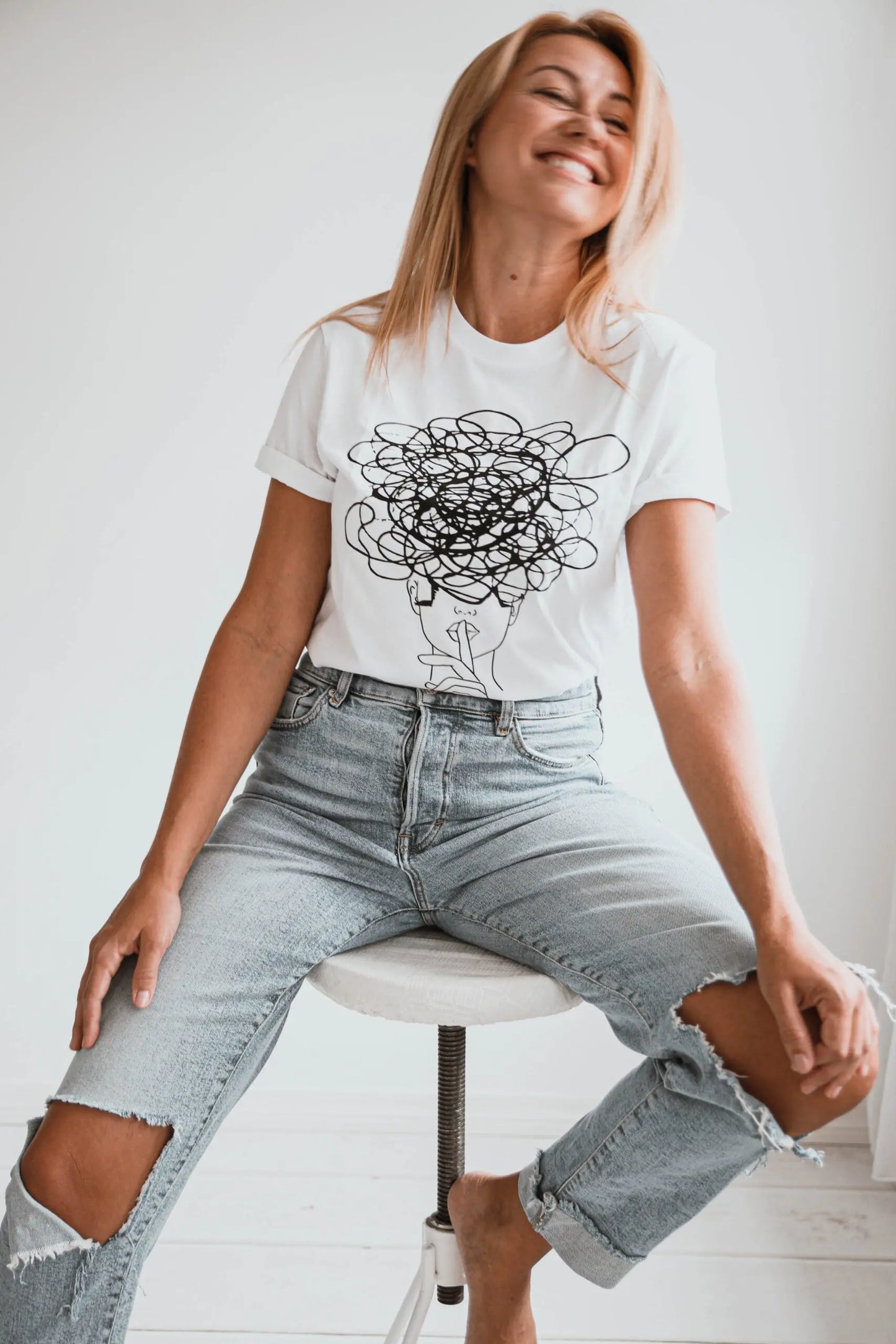 A woman in jeans sits on a stool, wearing a casual Ssshhh! T-shirt. Product features an oversize fit. Size chart: XS - XL. Washing instructions included.