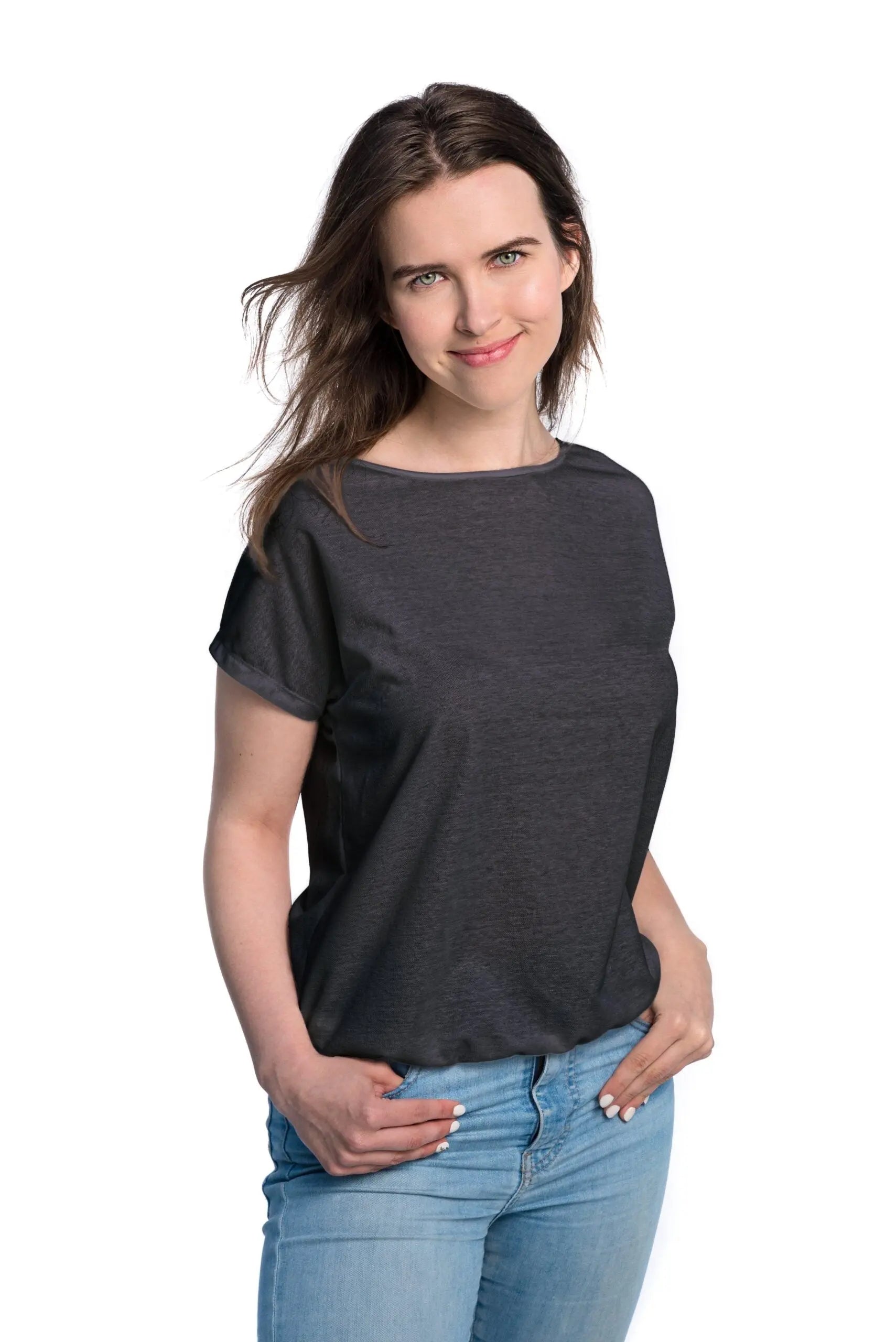A woman in a black shirt with hands in pockets, showcasing a Tan-Through T-Shirt - Dark Grey. Spacious, elastic-bottomed shirt for outdoor activities, allowing healthy tanning with unique fabric blend. Sizes XS-XXL available.