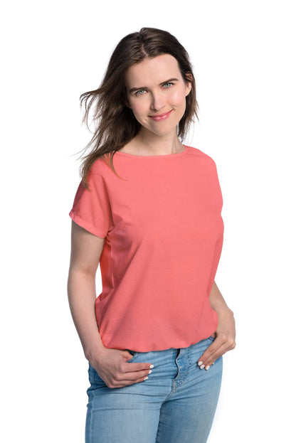 A woman in a coral tan-through T-shirt with hands in pockets. Spacious cut for outdoor comfort, elastic band at bottom, fabric allows UVA rays, 60% cotton, 40% polyester blend for quick drying.