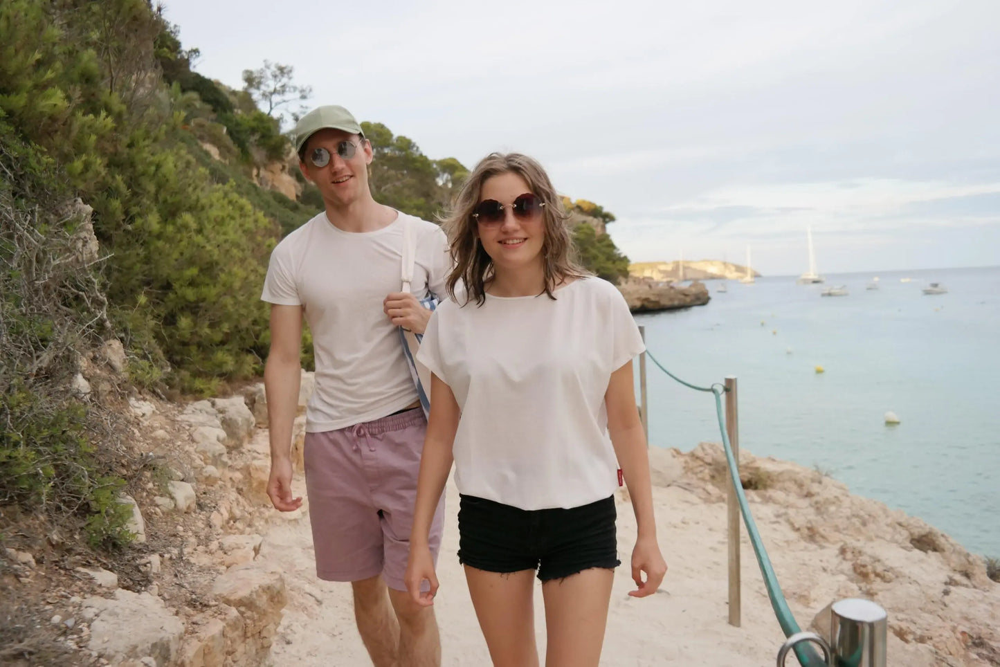 A man and woman in white shirts walk near water, wearing tan-through coral spacious T-shirts for outdoor activities. The fabric allows healthy tanning with UVA ray penetration. Sizes XS-XXL available.