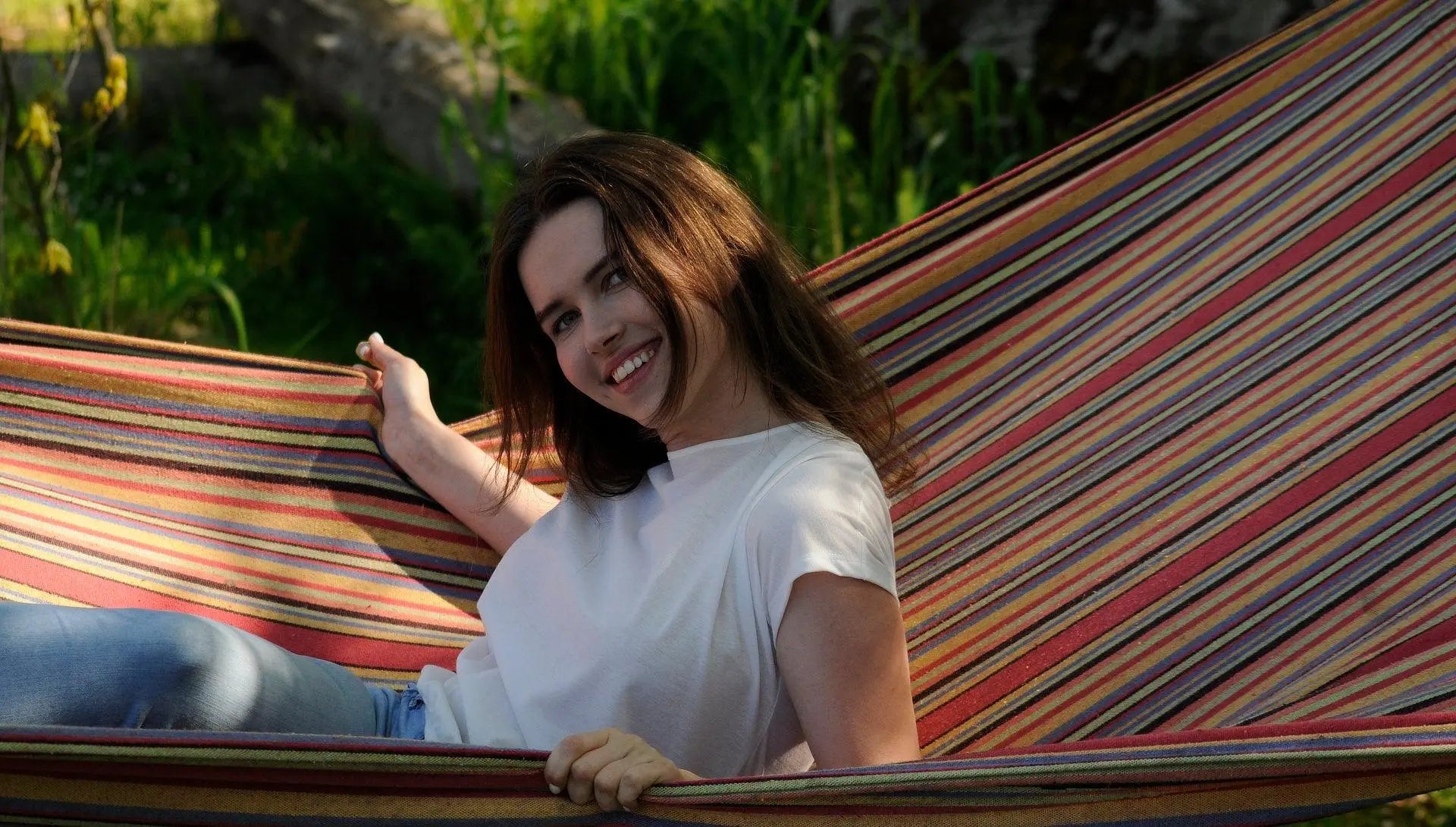 A woman in a hammock, smiling, wearing a Tan-Through T-Shirt in Coral. Spacious cut for comfort, elastic band at the bottom. Fabric allows healthy tanning with UVA penetration. Sizes: XS-XXL.