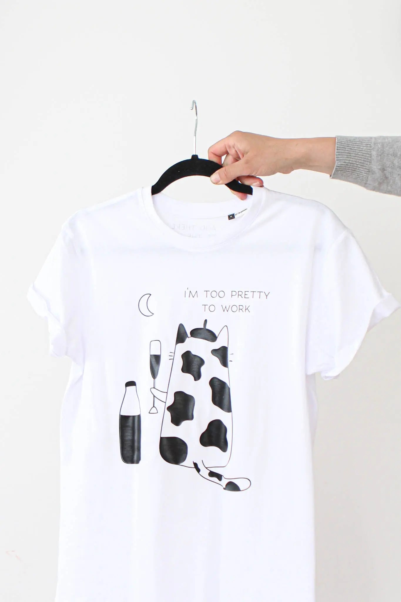 A hand holds an oversized white t-shirt with a cow print. Text reads I'm too pretty to work. Organic cotton. Size chart: XS to XL. Washing instructions included.