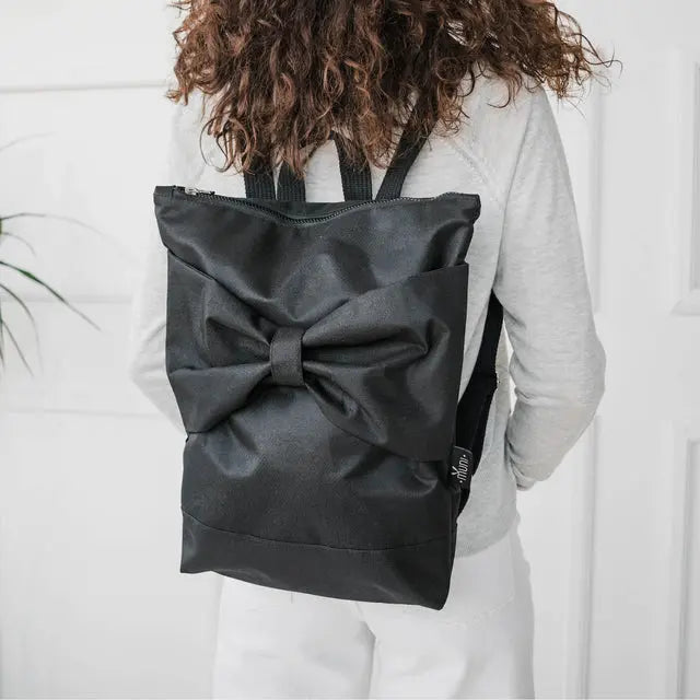 A waterproof Black Bow Backpack with a large bow detail. Versatile and durable for work or play. Features adjustable straps, zipper closure, and pockets for a 13 laptop. Dimensions: 38cm x 34cm.