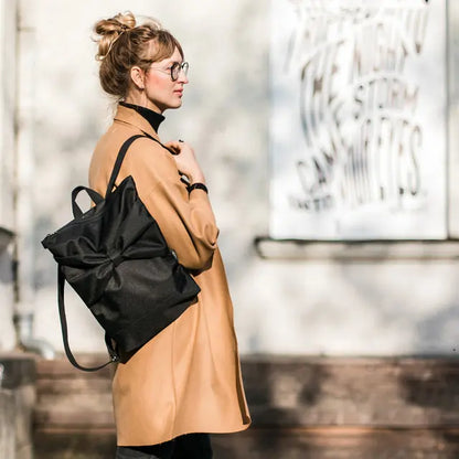 A woman in a brown coat carries a Black Bow Backpack, featuring waterproof black polyester with a large bow. Versatile and durable for work or leisure, with adjustable straps and laptop pocket.