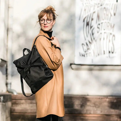 A woman in a brown coat carrying a Black Bow Backpack. Waterproof, with adjustable straps, zipper closure, and laptop pocket. Versatile and cute for work or outings.