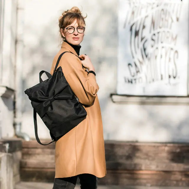 A woman in a brown coat wears a Black Bow Backpack. Waterproof and versatile, with adjustable straps and a laptop pocket. Dimensions: 38cm height, 34cm width.